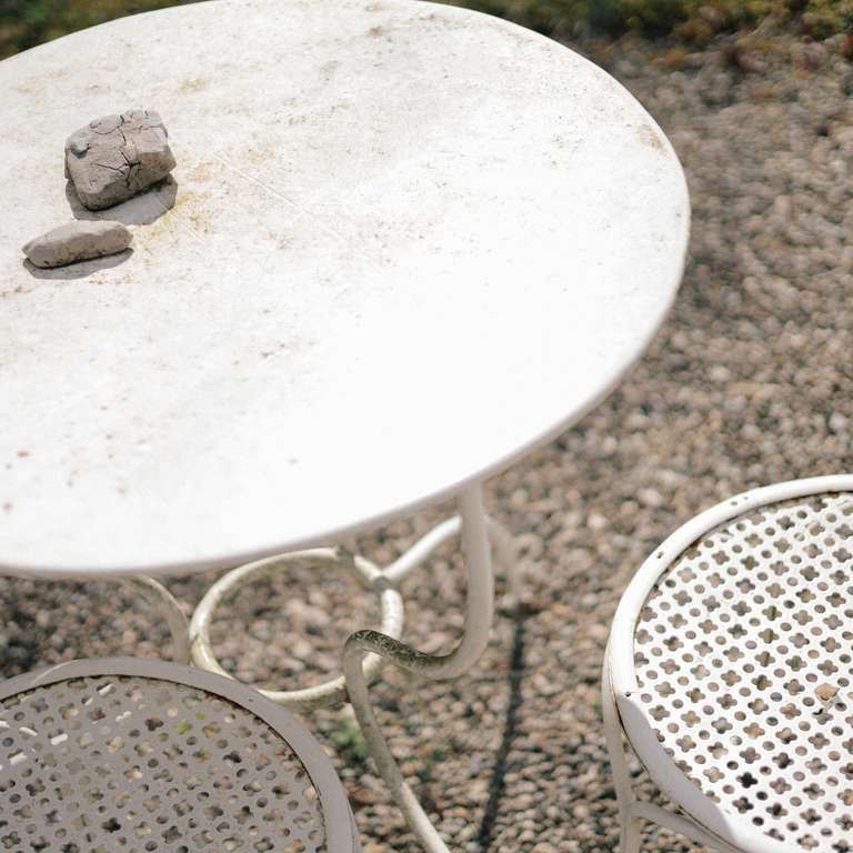 A round white metal table with two stones on top, next to a matching metal chair, sits on a gravel surface outdoors.